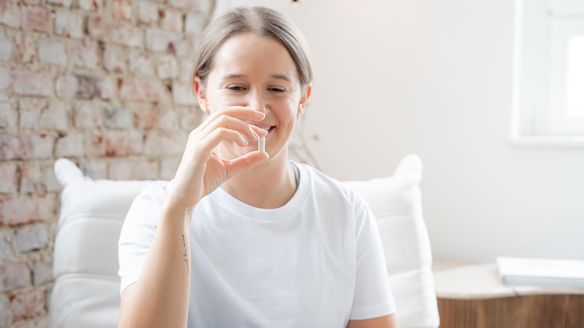 Junge Frau sitzt auf einem Sessel, lächelt und schaut auf Ihre Hand in der sie eine Kapsel Nahrungsergänzungsmittel für Frauen zwischen den Fingern hält