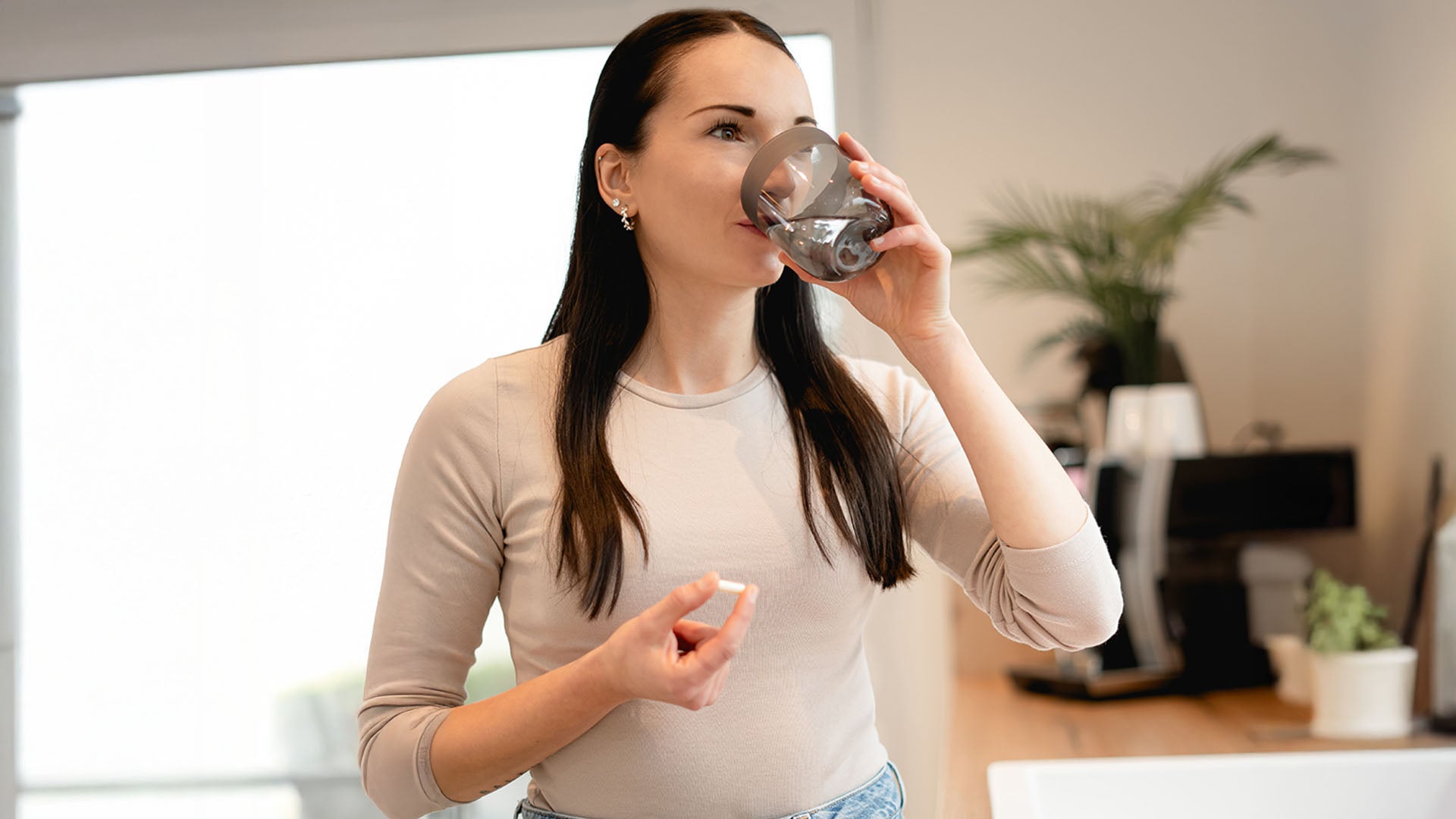eine frau trinkt aus einem glas wasser und hält in der anderen hand eine kapsel nahrungsergänzungsmittel